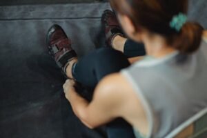 A female climber adjusts her shoes while sitting on a mat in a gym. Climbing shoes